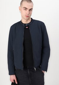 Modra bomber jakna iz lahkega materiala z okroglo ovratnikom, sprednjim zadrgo in dvema stranskima žepoma; oblečena čez črno srajco.