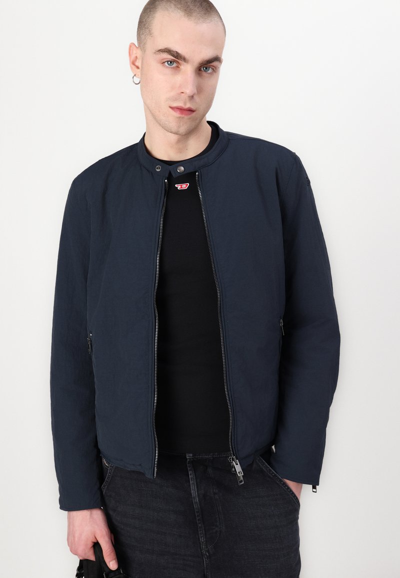 Veste bombardier bleu marine en tissu léger avec un col rond, zip sur le devant et deux poches latérales ; portée sur une chemise noire.