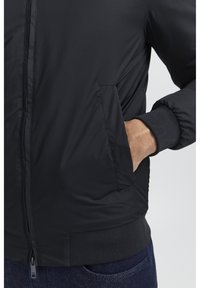Giacca nera isolata con una texture liscia, dotata di tasche laterali, chiusura con zip e polsini e orlo a costine.