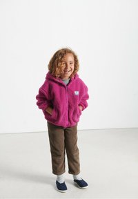 Lille barn med krøllet hår iført en lys pink zip-hoodie, brune kordfløjsbukser, stribede sokker og marineblå slip-on sko, som smiler.