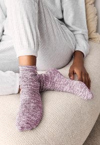 Paarse en witte gebreide sokken met een gestructureerd patroon, gedragen aan een voet. Zacht materiaal en een nonchalant ontwerp, geplaatst tegen een lichte, gestructureerde achtergrond.