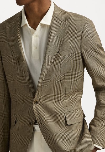 Blazer texturat în carouri maro, cu rever în formă de notch, închidere cu două butoane și buzunar pe piept, asortat cu un tricou polo alb dedesubt.