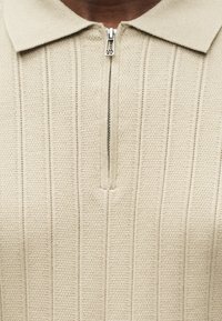 Polo shirt beige à fermeture éclair avec un motif de tricot texturé et des rayures verticales. Il possède un col et un tirette en métal.