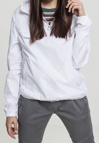 Persona che indossa un windbreaker bianco con cappuccio sopra una maglietta a righe, con pantaloni grigi, in piedi con una mano vicino al viso e l'altra rilassata lungo il corpo.