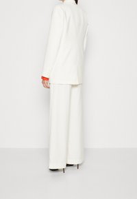 Vit skräddarsydd blazer med långa ärmar, kombinerad med vida byxor. Röda manschetter synliga. Slät textur, minimalistisk design och smal passform.