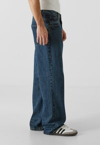 Mørkeblå denimjeans med løs passform, fem lommer, synlige sømmer og en lett falming. Kombinert med svarte og hvite sneakers.