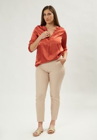 Blusa in satin arancione bruciato con scollo a V e dettagli arricciati, abbinata a pantaloni slim beige e sandali dorati a scivolo. Sfondo neutro.