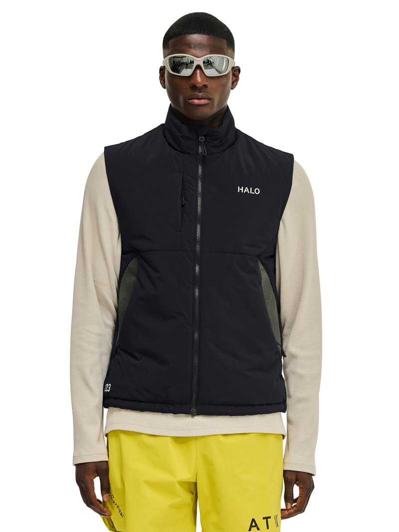 Mężczyzna ubrany w białą koszulę z długim rękawem, czarną bezrękawnik na zamek błyskawiczny z napisem "HALO", żółte spodnie i białe sportowe okulary przeciwsłoneczne, stojący na białym tle.