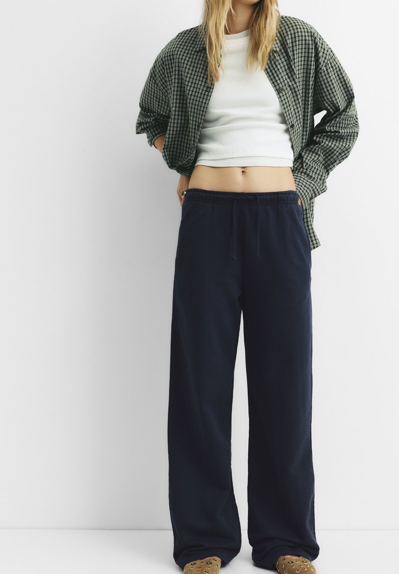 Weite marineblaue Hosen mit einem elastischen Bund, kombiniert mit einem weißen, enganliegenden Oberteil und einem oversized, grünkarierten Hemd, vor einem schlichten Hintergrund.