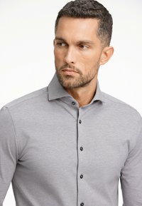 Camicia grigia con bottoni realizzata in un tessuto liscio, con colletto classico e bottoni neri lungo la pattina anteriore.