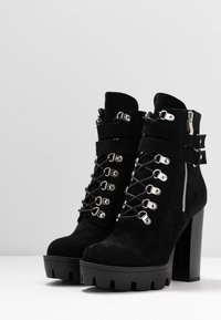 Stivaletti alla caviglia in suede nero con tacco spesso, dotati di accessori in metallo argentato, lacci sul davanti e cerniera laterale. Suola in gomma larga con bordo seghettato.