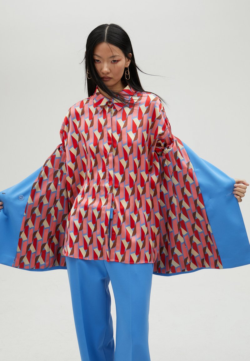 Patterned red and blue shirt featuring geometric shapes, made of shiny fabric. Paired with solid blue wide-leg pants.