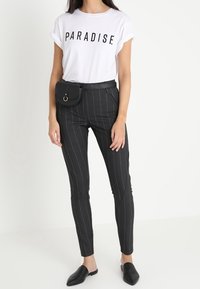 Žena ve světle bílé tričku s nápisem "PARADISE", černých kalhotách s proužky, černé ledvince a černých nazouvacích botách, stojící.