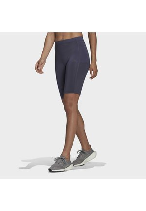Femme portant un short de sport gris foncé mi-cuisse et des chaussures de course grises, debout, jambes croisées, sur un fond clair uni.