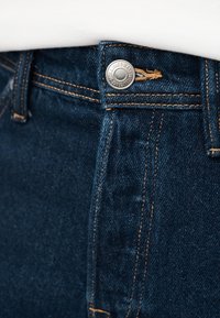 Κοντινή λήψη μέσης σκούρων μπλε τζιν με μεταλλικό κουμπί και αντίθετη ραφή πορτοκαλί χρώματος.