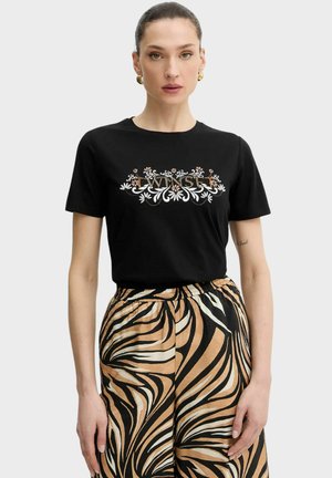 Donna che indossa una maglietta nera a maniche corte con il testo floreale "ENSE" e pantaloni a vita alta con strisce zebrate beige, nere e bianche.