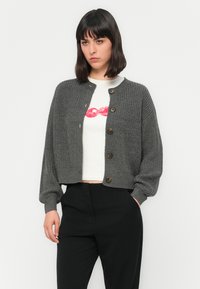 Una donna con capelli scuri di media lunghezza indossa un cardigan grigio con bottoni sopra un crop top bianco con una grafica di labbra rosse e pantaloni neri.