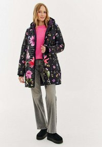 Černý vycpávaný kabát s květinovým potiskem, zapínáním na zip a kapucí. Nošený přes růžový svetr a šedé džíny s černými botami na platformě.