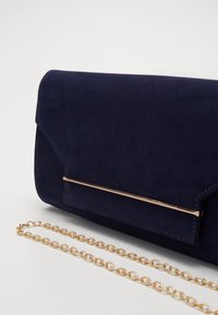 Clutch in suede blu navy con dettaglio in barra dorata e tracolla coordinata in oro su una superficie bianca.