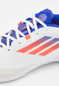 Zapatilla deportiva blanca con un diseño texturizado, acentos azules y franjas de color naranja brillante. Presenta cordones, material sintético y un cuello acolchado.