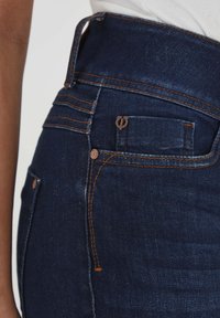 Sötétkék, magas derekú farmernadrág narancssárga varrással, hátsó zsebbel és sima denim textúrával. Derékrészen övbújtató található.