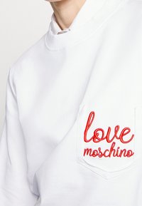 Vit sweatshirt med ribbad halsringning, med röd broderad text "love moschino" på vänster bröstficka. Slät textur.