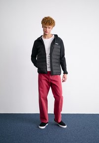 Černá vycpaná bunda s kapucí, nošena přes bílou košili. Spárováno s volnými červenými kalhotami a černými slip-on botami. Jednoduchý design a pohodlný střih.