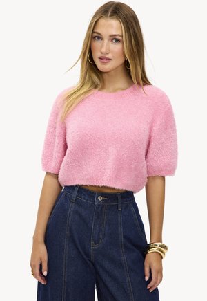 Suéter corto de manga corta en color rosa con textura suave, y un escote redondo. Combinado con pantalones vaqueros oscuros de tiro alto y pulseras doradas.