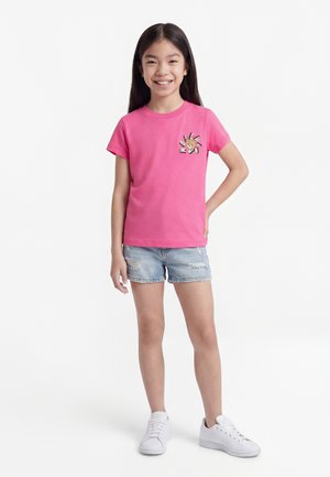 Κορίτσι με μακριά σκούρα μαλλιά, που φοράει ροζ μπλουζάκι με στάμπα, ανοιχτό μπλε σορτς από τζιν και λευκά αθλητικά παπούτσια, χαμογελώντας με το ένα χέρι στη μέση.