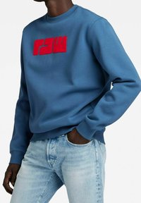 Sudadera azul con cuello redondo, con un gran logotipo rojo "RAW" en el pecho, hecha de una tela suave. Llevada con jeans de un azul claro.