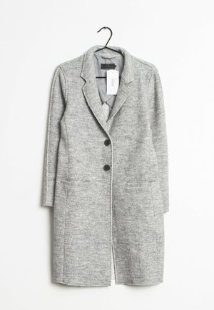 Manteau long en laine gris clair avec revers, deux boutons noirs, poches avant, suspendu à un cintre noir avec une étiquette de prix blanche.
