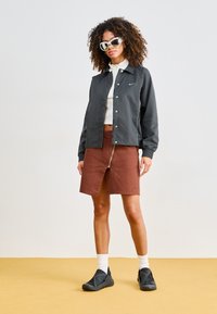 Juodas lengvas švarkas su spaudėmis, baltas aukštu kaklu viršus, rudai spalvos džinsinė sijonas su užtrauktuku, ir juodi batus be užtrauktuko su baltais kojinaitėmis.