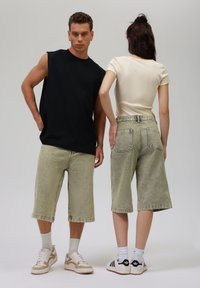 Black sleeveless top, cream ribbed shirt, green denim shorts. White and black sneakers. Visible back pockets and stitching details.