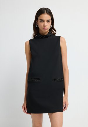 Mujer con un vestido mini negro sin mangas, con un escote alto de cuentas y detalles de bolsillos también de cuentas, de pie frente a un fondo blanco.