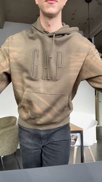 Personne portant un sweat à capuche GAP marron et un pantalon sombre, debout dans une pièce avec des chaises et une table visibles en arrière-plan.