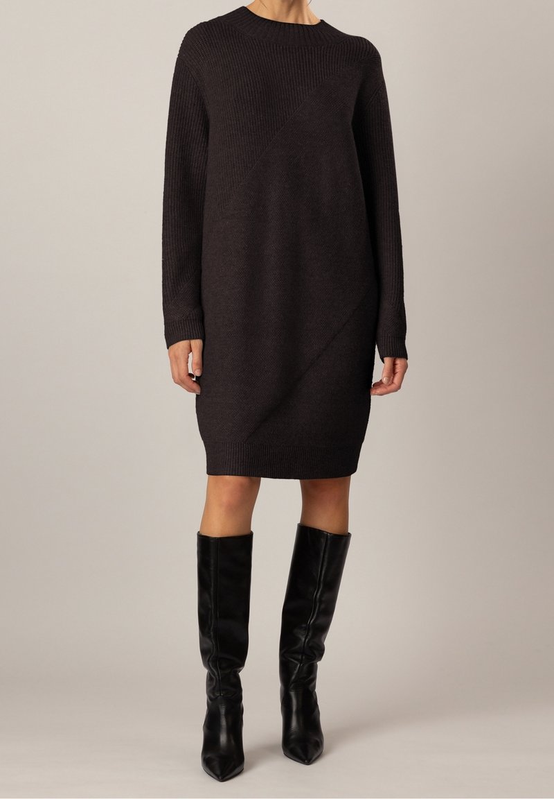 Femme portant une robe en maille côtelée marron foncé à manches longues et longueur genou, ainsi que des bottes en cuir noires montantes, debout devant un fond uni.