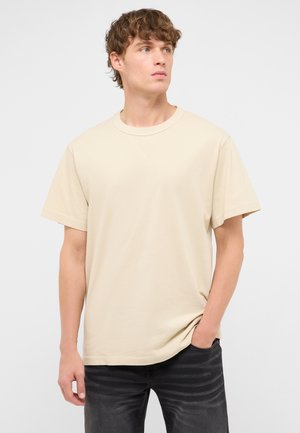 Młody mężczyzna z kręconymi włosami, ubrany w beżowy t-shirt z krótkim rękawem i czarne dżinsy, stoi z jedną ręką w kieszeni, patrząc na bok.