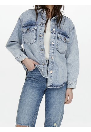 Frau trägt eine hellblaue Jeansjacke über einem weißen Oberteil und zerrissenen blauen Jeansshorts, mit der Hand in der Tasche vor weißem Hintergrund.