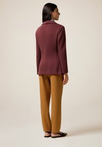 Cappotto bordeaux con design sartoriale e colletto a punte, abbinato a pantaloni dritti color senape. Tessuto morbido, dettagli minimali.