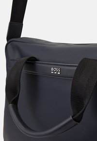 Bolso azul marino con una superficie texturizada, que presenta una correa negra y un mango liso. Un logo plateado de "BOSS" se muestra en el panel frontal.