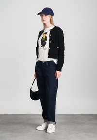 Cardigan preto de malha com botões dourados, t-shirt gráfica branca, jeans escuros e ténis brancos. Acessório: mala preta, boné azul-marinho.