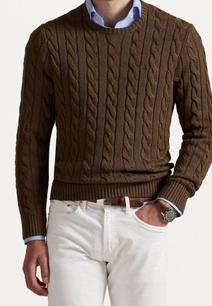 Man draagt een bruine kabelgebreide trui over een blauw overhemd met kraag, witte broek, bruine riem en polshorloge, met één hand in de zak.