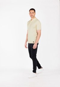Beżowa koszulka polo z krótkim rękawem, wykonana z gładkiego materiału, w połączeniu z czarnymi slim-fit dżinsami i białymi trampkami, stojąca na jasnym tle.