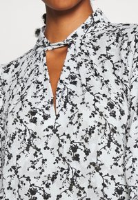 Blusa in tessuto azzurro chiaro con stampa floreale nera, dotata di un'apertura a occhiello e di un colletto con un dettaglio di cravatta applicato.