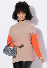 Maglione beige lavorato a maglia con colletto alto e maniche arancioni vivaci, abbinato a una borsa grigia luccicante. Presenta orli a coste e aperture laterali.