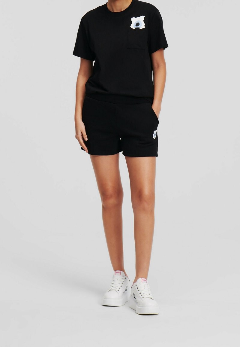 Femme portant un short noir et un t-shirt noir avec un petit motif blanc sur la poche poitrine et le short, assortis à des baskets blanches.
