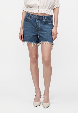 Femme portant un short en jean bleu effiloché, un chemisier blanc court et des chaussures à talons beiges, debout devant un fond uni.