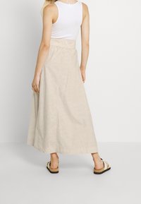 Beige maxi kjol med ett texturerat mönster, elastisk midja och sidofickor, kombinerad med platta sandaler med korksula.