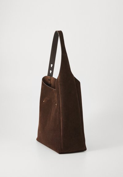 Sac fourre-tout en suède marron avec un côté plat, une forme structurée et une courte poignée en cuir marron. Présente de petites clous dorés sur le devant.