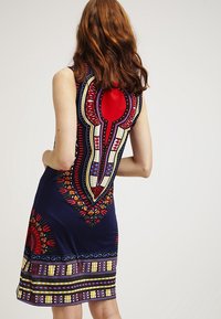 Granatowa sukienka z kolorowymi, misternymi wzorami w odcieniach czerwieni, żółci i niebieskiego. Posiada wycięcie na plecach w kształcie klucza oraz dopasowany krój.
